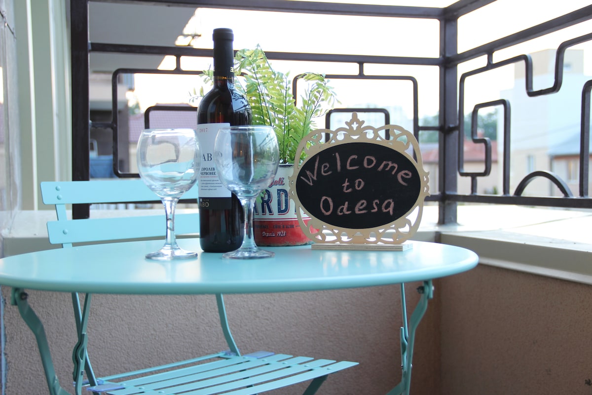A small balcony table is set with two wine glasses and a bottle of red wine, accompanied by a decorative plant. A sign reading 'Welcome to Odesa' adds a personal touch to the inviting outdoor space.