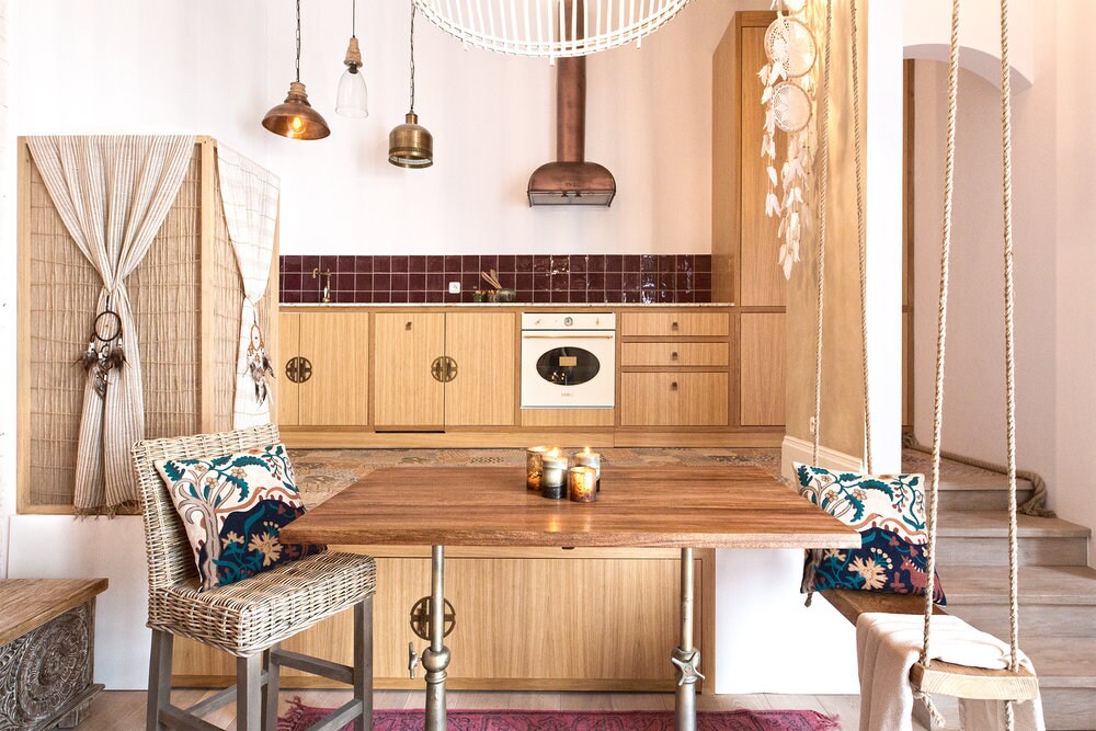 A wooden dining table is centered in the open space, accompanied by two woven chairs adorned with decorative cushions. A kitchen area features wooden cabinetry and a distinctive hood above the stove, complemented by warm lighting fixtures and a cozy atmosphere.