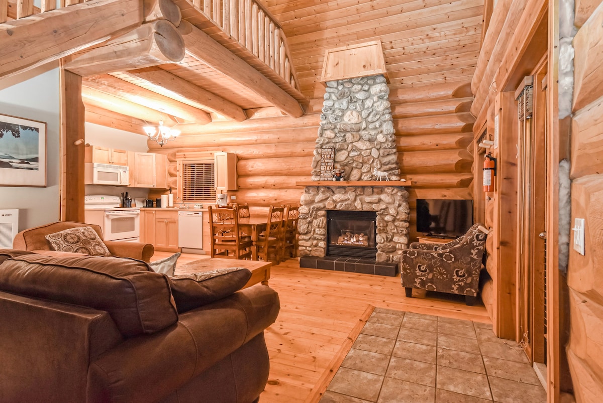 An inviting living area features a stone fireplace that adds warmth to the space. A spacious couch and patterned armchair provide comfortable seating, with wooden elements throughout. The open kitchen with modern appliances and a dining table is visible, creating a welcoming atmosphere for relaxation and meals.