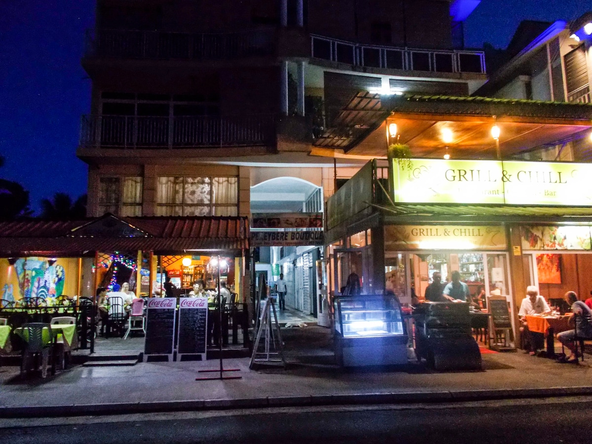 The image captures a vibrant street scene at night featuring two restaurants, one illuminated with bright signage. Colorful lights adorn the outdoor seating area, and patrons are visible enjoying their meals. The building's architecture includes balconies and a covered walkway, enhancing the inviting atmosphere.
