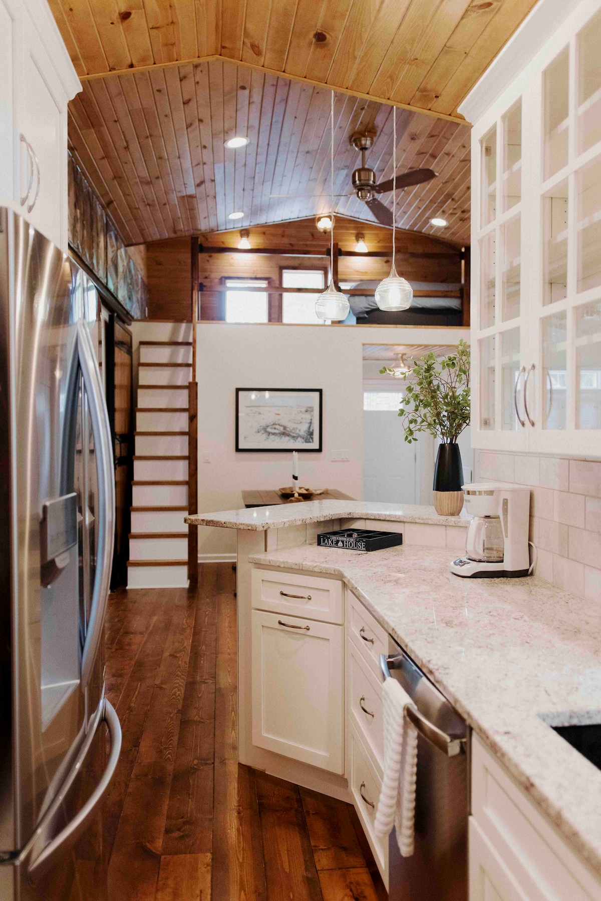 The kitchen features a granite countertop paired with white cabinetry, complemented by stainless steel appliances. The high, wooden ceiling enhances the spacious feel. A staircase leads to the loft area, while natural light filters through nearby windows, highlighting the modern design.