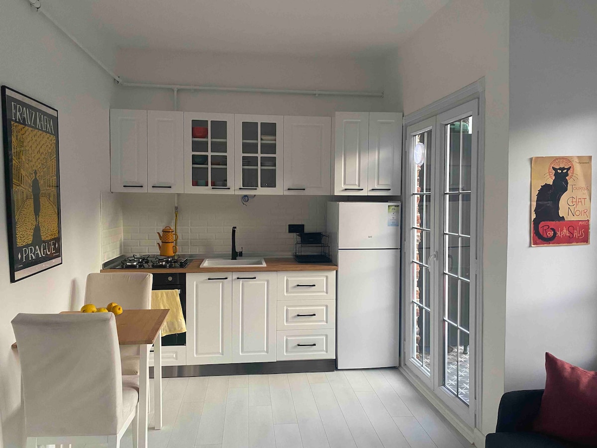 A modern kitchen area showcases white cabinetry and a wooden countertop. A dining table for two is illuminated by natural light from the glass doors. An appliance suite is neatly arranged, with a refrigerator and a gas stove complementing the light-colored decor.