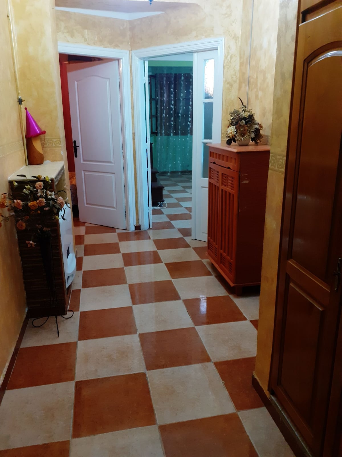 A corridor is presented, featuring a checkered floor with alternating brown and cream tiles. Doors to multiple rooms are visible along the walls. Decorative items, including a vase and small flowers, are positioned on a console table. Light-colored walls enhance the space's brightness.