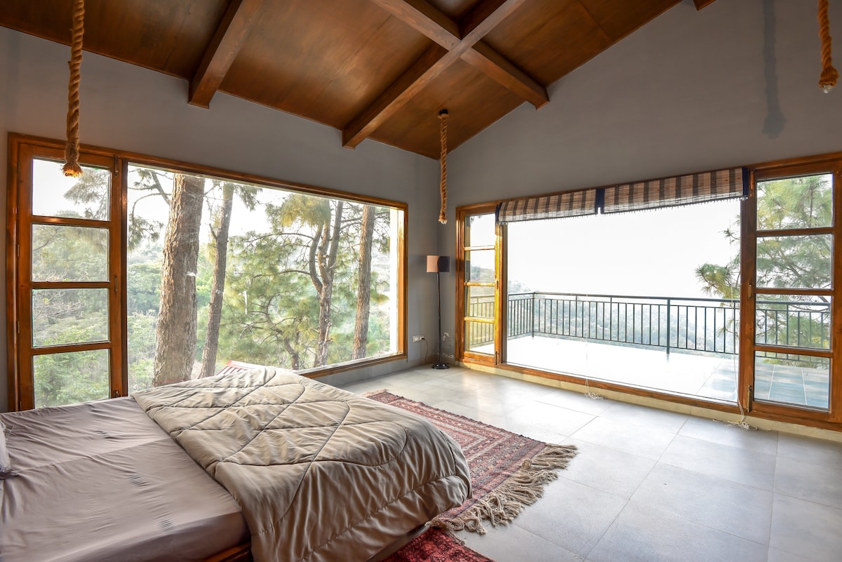 A spacious bedroom features floor-to-ceiling windows that offer views of the surrounding trees and mountains. A minimalist bed is complemented by a patterned rug beneath. Natural light fills the room, enhancing the calming atmosphere of the uncluttered space.