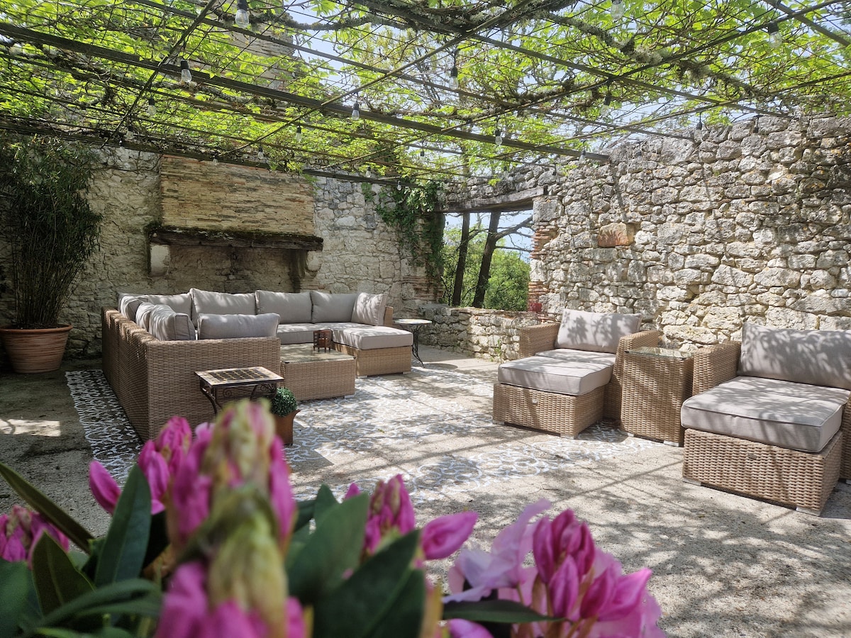 An outdoor seating area is framed by lush greenery and rustic stone walls. Comfortable wicker furniture is arranged in a relaxed layout, complemented by decorative pillows. A patterned rug adds warmth to the space, which is shaded by a vine-covered pergola, providing a serene setting for gatherings.