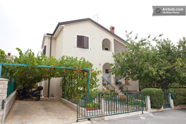 A two-story residential building is surrounded by lush greenery, featuring a covered parking area. The entrance is highlighted by a set of stairs leading to the front door, with decorative plants situated alongside the pathway.
