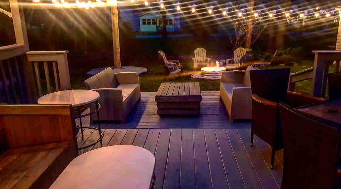 A cozy backyard space is revealed, featuring a lighted lounge area with comfortable couches and a low table. Adirondack chairs surround a fire pit, creating a welcoming spot for relaxation. Warm string lights hang overhead, enhancing the atmosphere during evening gatherings.