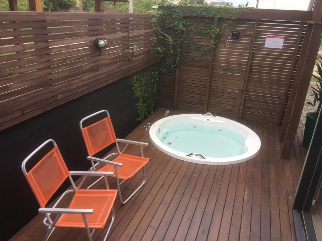 An outdoor area features a private jacuzzi surrounded by wooden fencing, offering an intimate setting. Two orange chairs are positioned beside the jacuzzi on a wooden deck, accented by greenery that adds a touch of nature to the space.