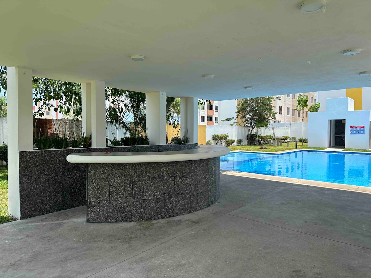 An outdoor space features a sleek bar area with a curved countertop, situated beside a swimming pool. The pool reflects surrounding greenery and buildings, while shaded seating areas are visible in the background, providing a relaxing atmosphere for gatherings.