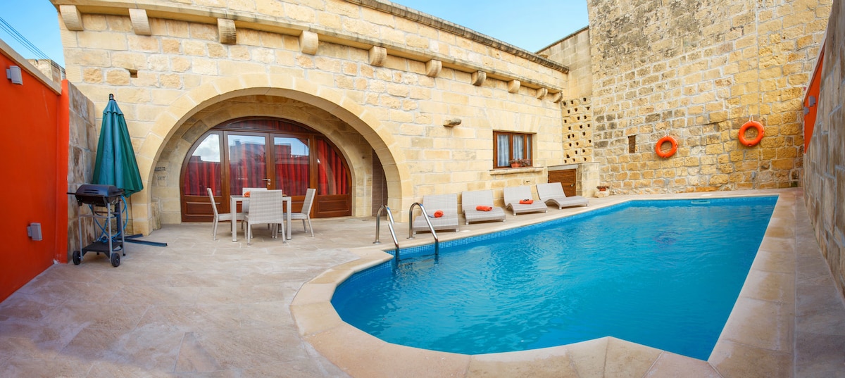 A private outdoor pool area is featured, surrounded by smooth stone walls. Lounge chairs are positioned near the water, with a small table and chairs for dining. A barbecue grill is placed nearby, and a shaded umbrella offers a respite from the sun.