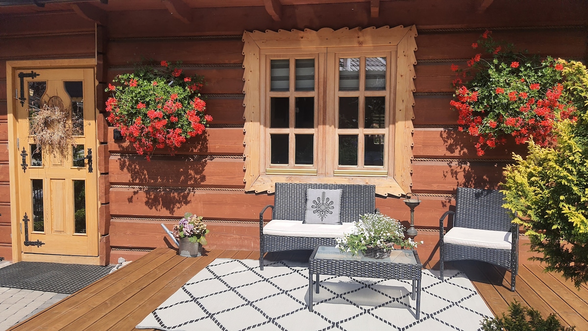 The exterior of the wooden house features a welcoming entrance with a decorative door and large window adorned with vibrant red flowers. Two comfortable seating areas with cushions flank a table, set on a patterned outdoor mat, creating an inviting space for relaxation.
