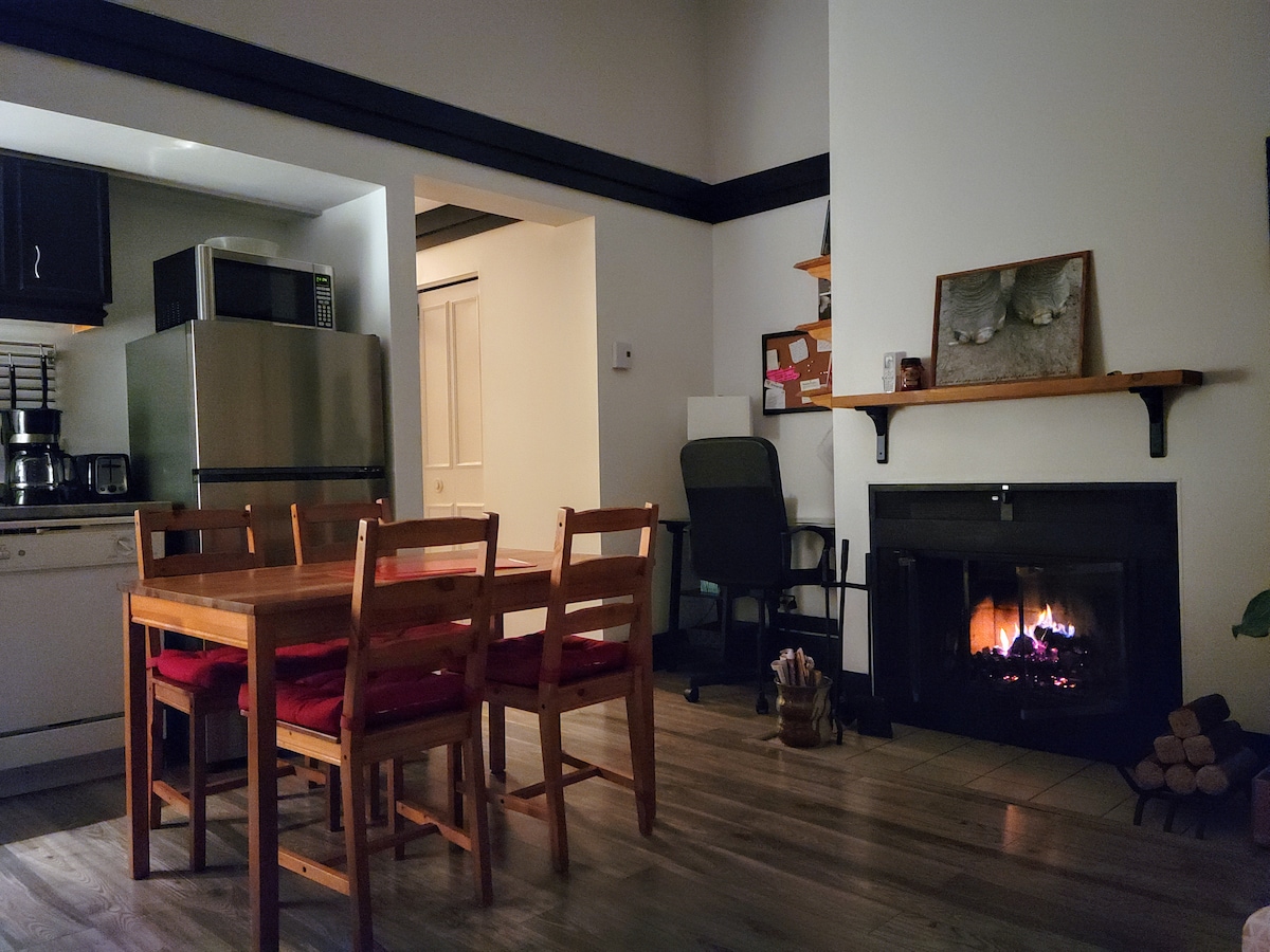 A dining area features a wooden table with chairs adorned with red cushions. A fireplace is situated nearby, flickering flames visible. In the background, a fully equipped kitchen is seen, along with a small workspace and shelves displaying decor items.