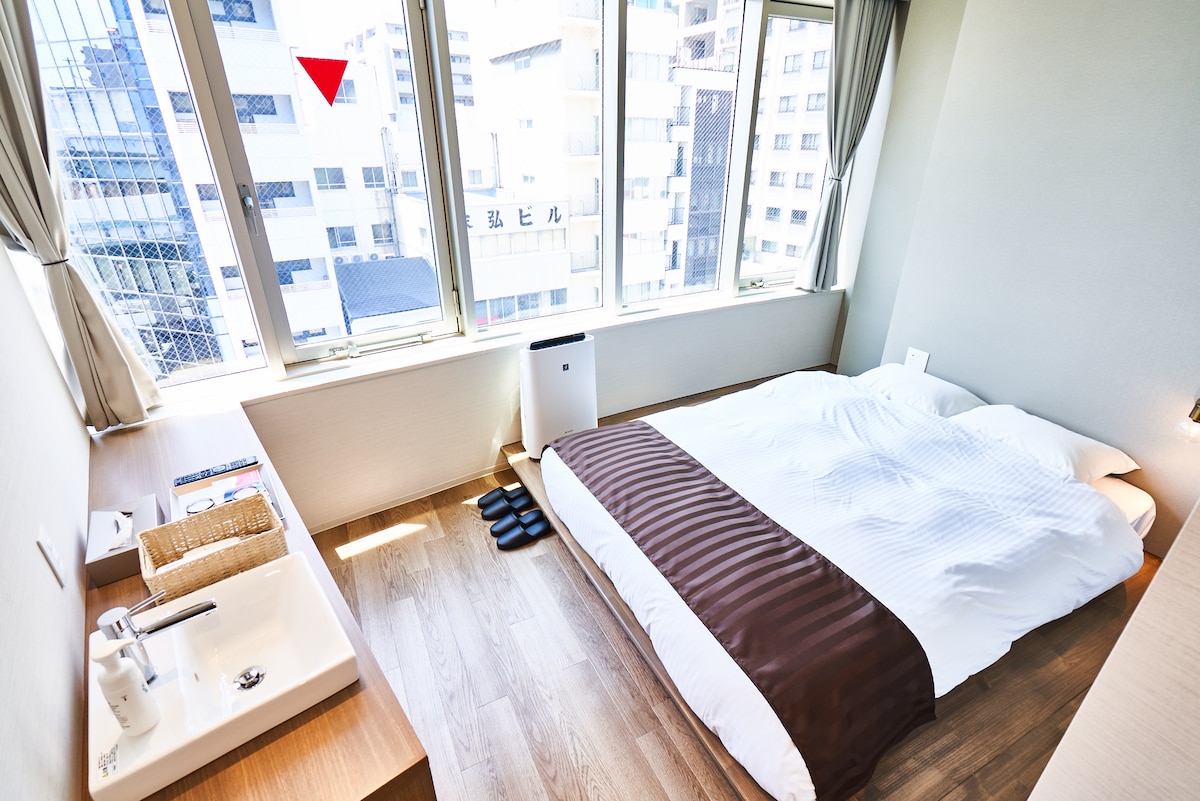 A bright room features large windows allowing natural light to fill the space. A spacious bed is positioned with a simple design. A small table and a pair of slippers are also visible. Nearby, a compact air purifier adds to the room's comfort.