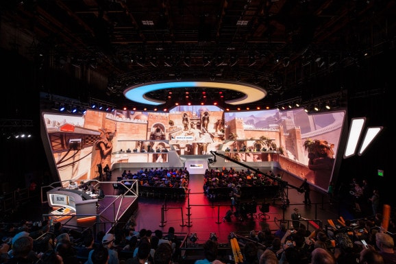 A large esports arena is shown, featuring a vivid stage adorned with massive screens displaying game graphics. Rows of audience seats are filled with spectators, while bright lighting illuminates the setup. The stage is equipped with gaming stations and cameras, creating an immersive atmosphere.