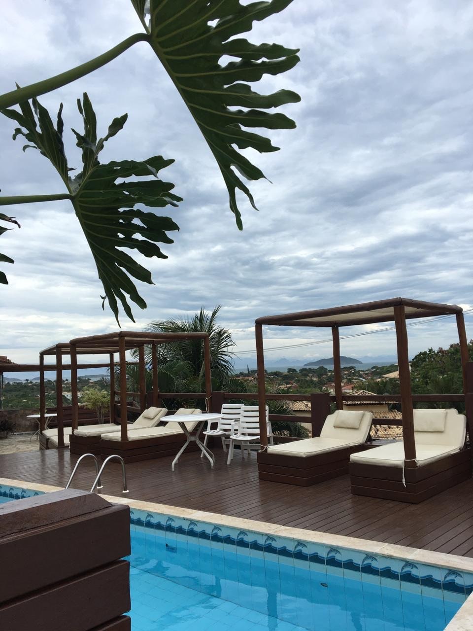 A serene pool area features a wooden deck with two sheltered lounge beds, accompanied by a dining table and chairs. Lush greenery and distant hills are visible, set against a cloudy sky, creating a relaxed atmosphere for leisure.