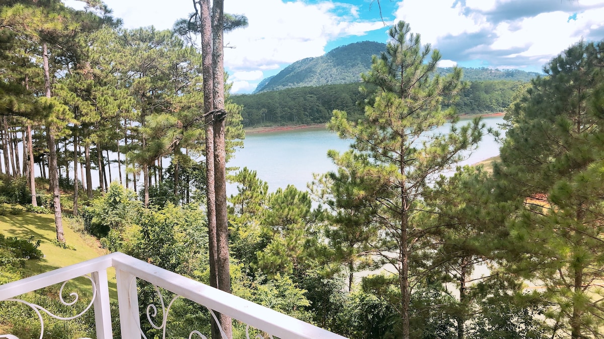 A serene view from the balcony reveals a tranquil lake framed by lush pine trees and rolling mountains. The landscape is bathed in natural light, highlighting the vibrant greenery and calm water that reflects the surrounding scenery.