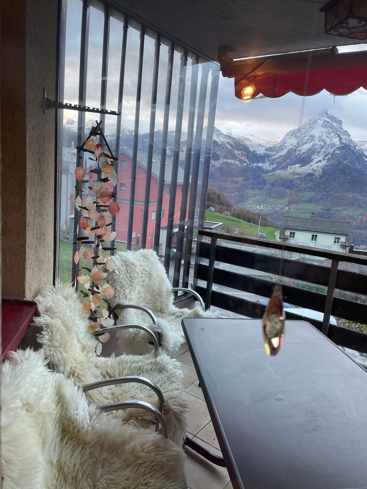 A covered balcony features a glass table surrounded by metal chairs, with plush white covers. A decorative hanging mobile adds color, while stunning mountain views are visible through the railing. The space is sheltered by an awning, creating a cozy outdoor atmosphere.