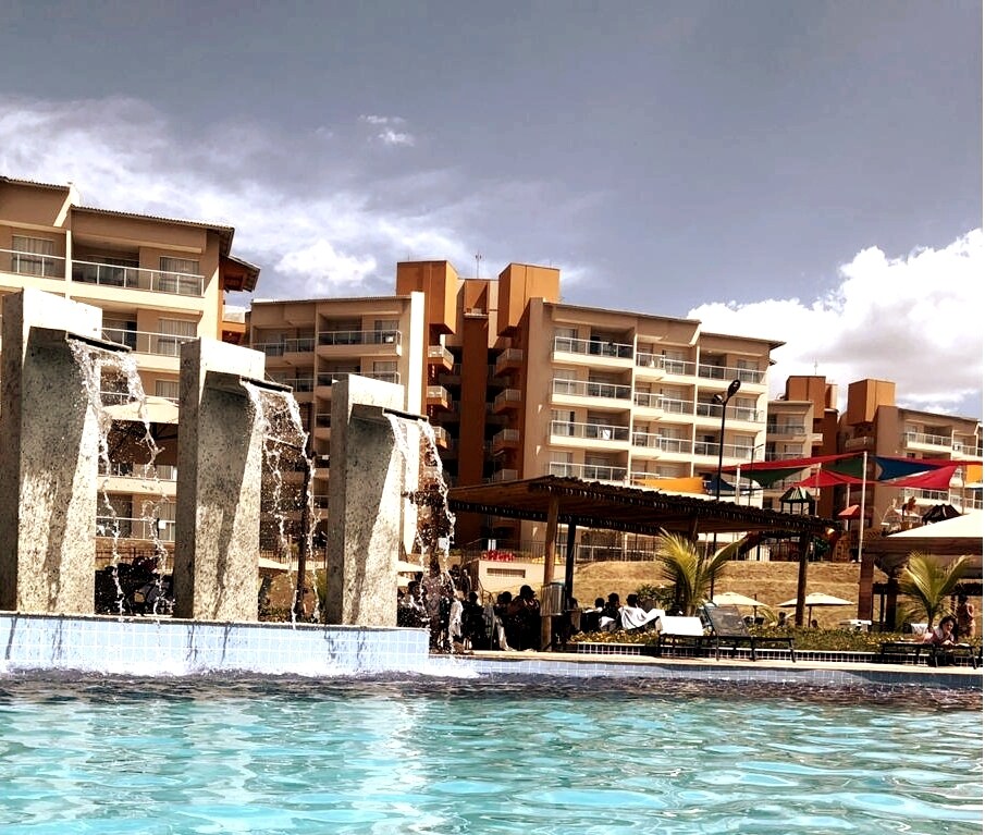 A vibrant pool area is highlighted by four sculptural water features cascading into the water. The backdrop features a multi-story resort building, with colorful canopies providing shade for seating areas, surrounded by greenery.