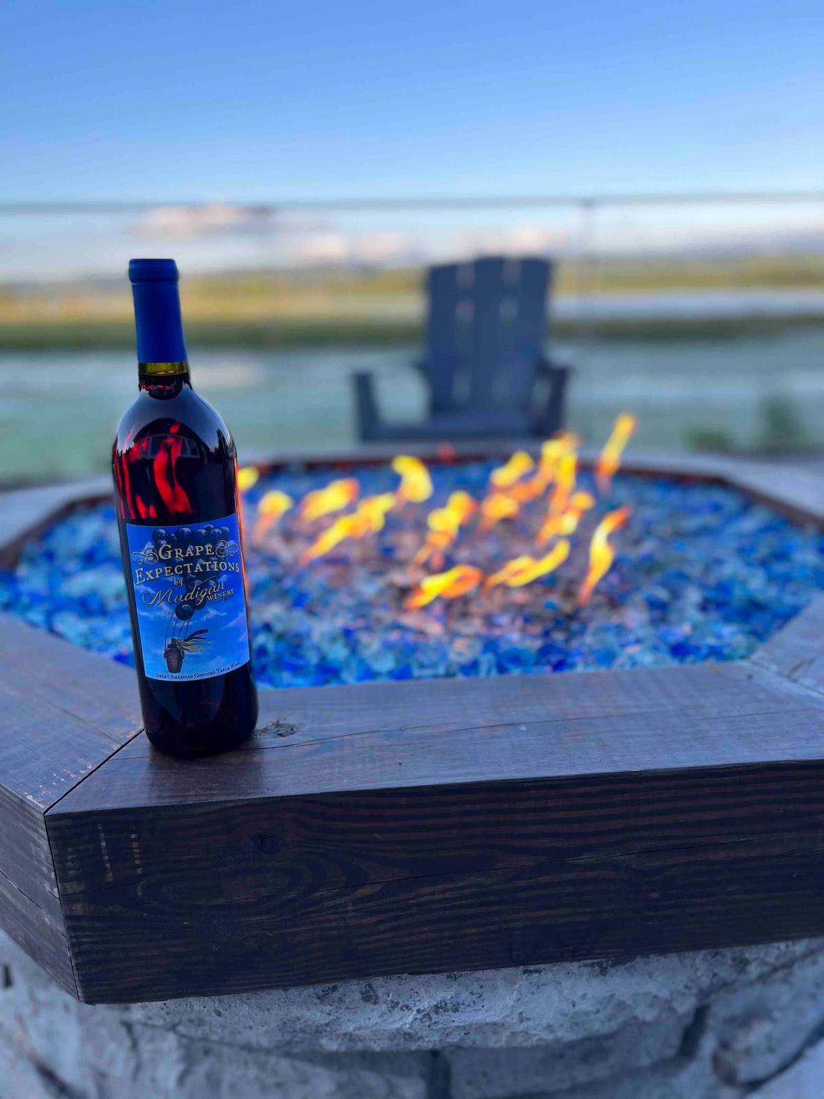 A bottle of red wine is positioned beside a fire pit, featuring vibrant blue glass stones. Flames dance within the stone enclosure, offering a warm glow against the backdrop of a tranquil outdoor setting. A single Adirondack chair is visible in the distance.
