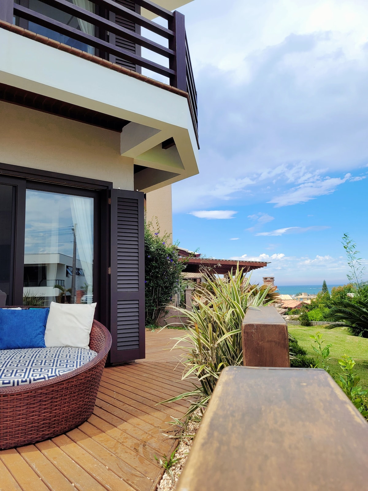 A welcoming outdoor seating area features a round, wicker chair adorned with cushions, situated on a wooden deck. Lush greenery and sea views are visible, while the sky displays a blend of clouds and blue in a serene atmosphere.