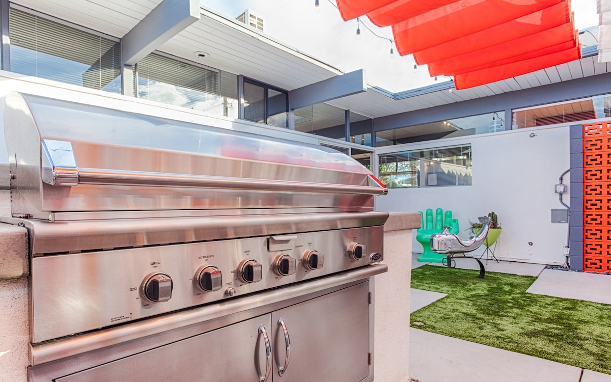 An outdoor kitchen area is equipped with a stainless steel grill, positioned against a backdrop of modern architecture. Colorful awnings provide shade over the space, and a vibrant green lawn is visible in the background, alongside lounge seating and decorative elements.