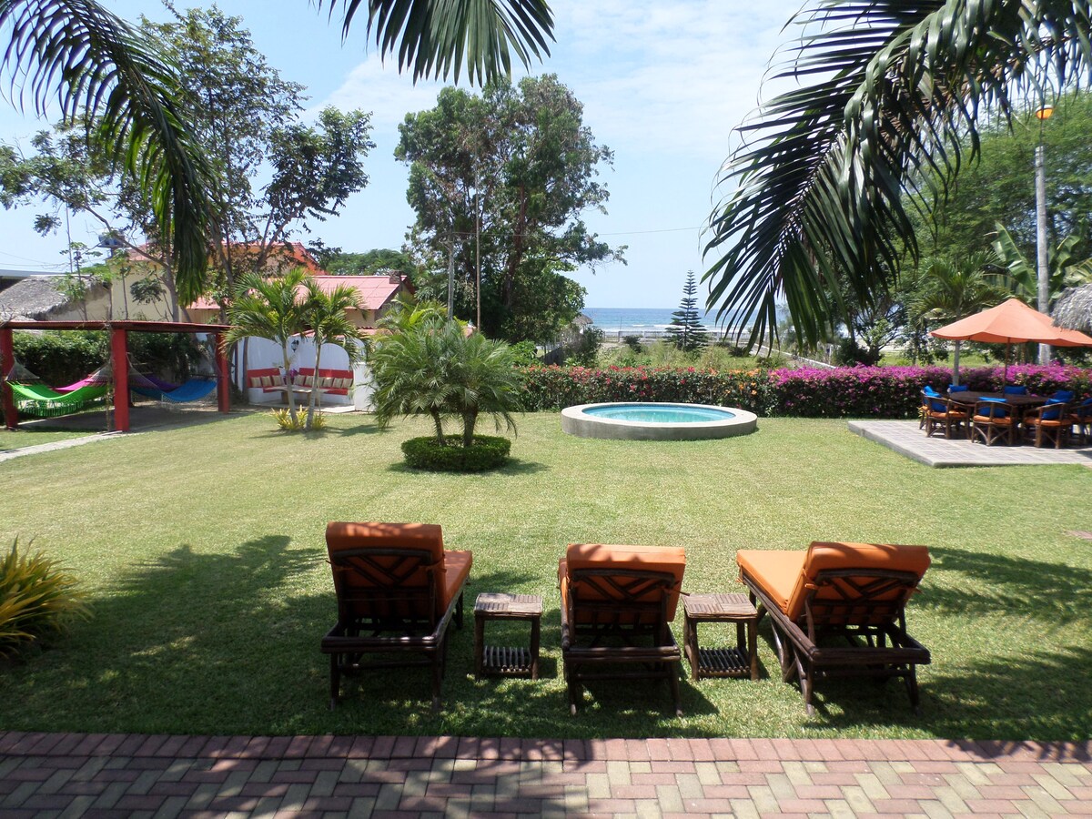 A serene outdoor area features well-manicured green grass, with three lounge chairs arranged invitingly under palm trees. A circular pool is visible nearby, surrounded by vibrant tropical plants. In the background, the ocean can be glimpsed, complementing the relaxing atmosphere.