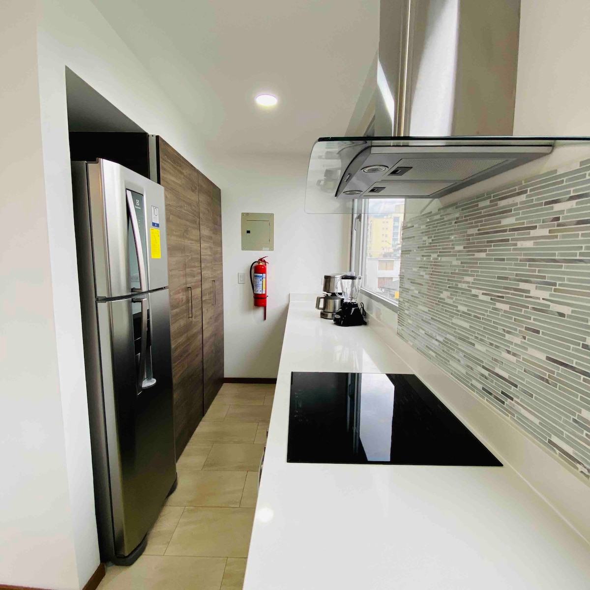 The kitchen features modern appliances, including a stainless steel refrigerator and a small coffee maker. A sleek induction cooktop is set into the smooth white countertop, with a contemporary tile backsplash providing a subtle textural contrast. Natural light brightens the space through the window.