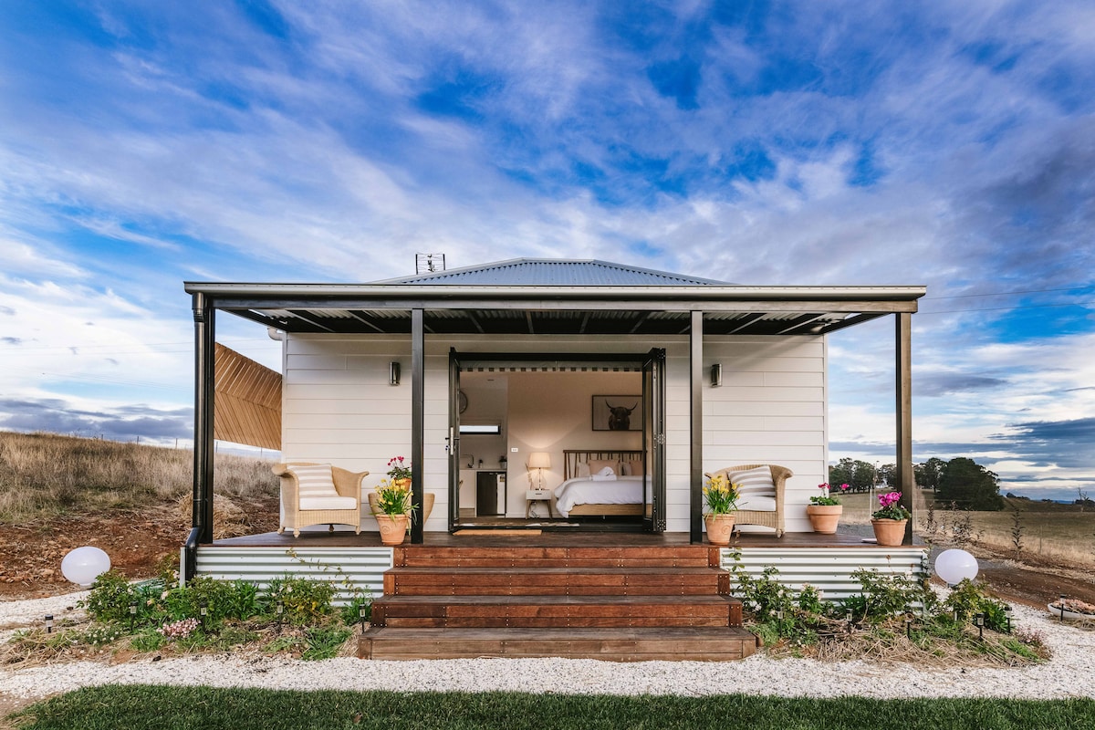A charming cottage features a welcoming exterior with wooden steps leading to a covered porch. Planters filled with flowers flank the entrance, while large glass doors reveal a bright interior. The backdrop showcases open fields and a dramatic sky, emphasizing the serene rural setting.