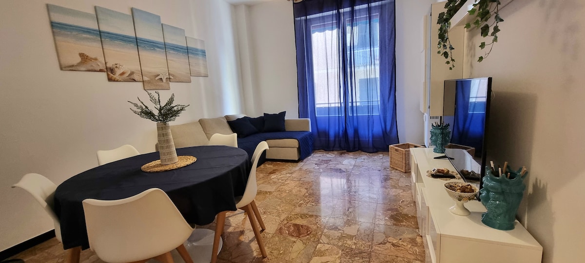 A spacious living area features a round dining table set for six. A comfortable sofa is positioned nearby, complemented by soft cushions. Large windows with flowing dark curtains allow natural light to enter, while beach-themed artwork adorns the wall, enhancing the coastal vibe.