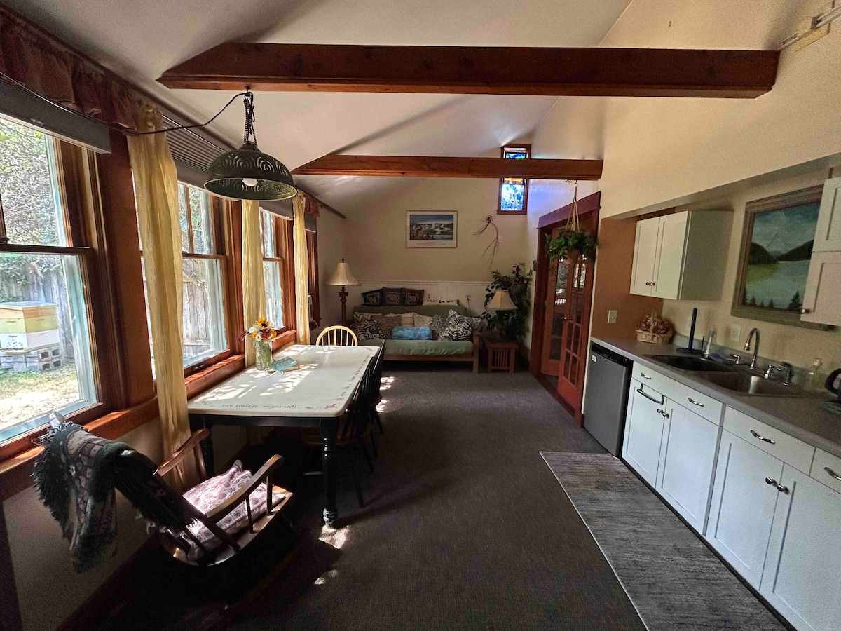 The interior space features a dining table surrounded by chairs, with a cozy seating area visible in the background. Natural light pours in through large windows, highlighting the vaulted ceiling and wooden beams. A kitchenette with essential appliances is located along one wall.