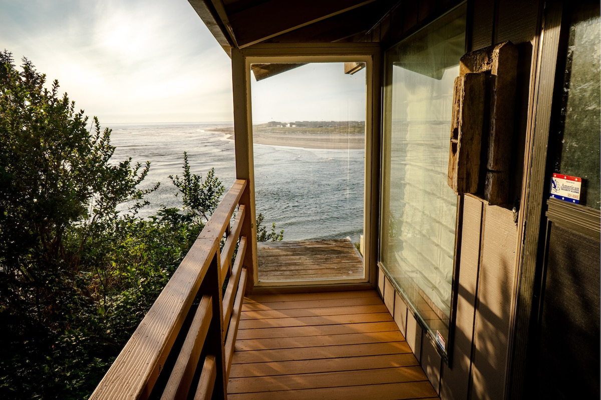 A spacious deck offers a panoramic view of the ocean and surrounding landscape. Natural light filters through large glass windows, creating a bright and airy atmosphere. Wooden railings frame the view, inviting guests to enjoy the serene coastal scenery.
