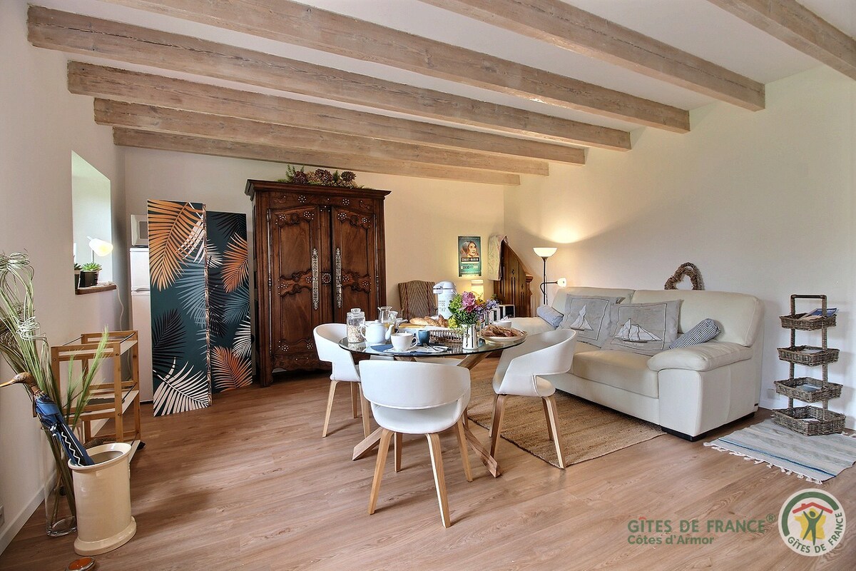 A private living area showcases a comfortable light-colored sofa and a wooden dining table set for guests. Four white chairs surround the table, while decorative elements include a dark wooden cabinet and wall art featuring tropical foliage. Exposed wooden beams enhance the open atmosphere.