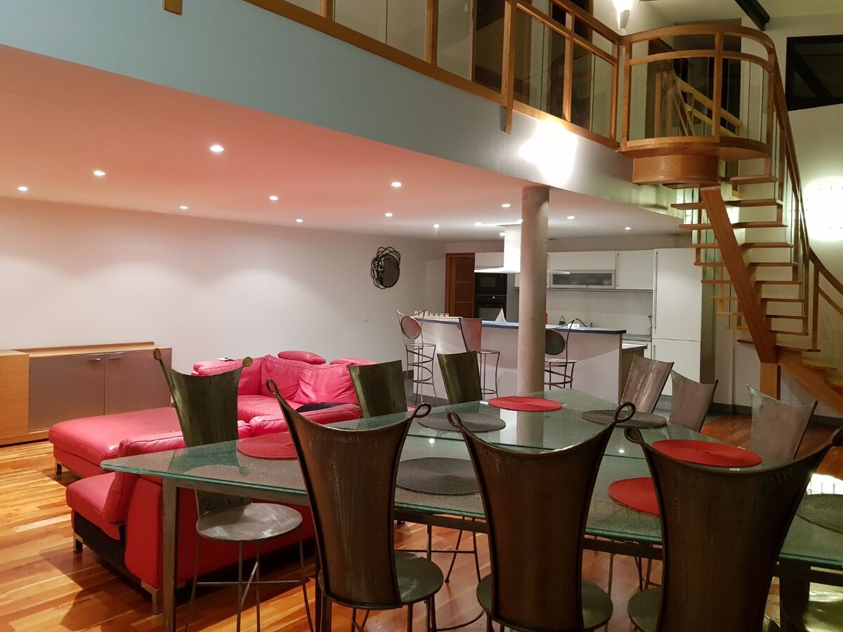An open dining area showcases a large glass table surrounded by several metal chairs, accented with red cushions. A modern kitchenette is visible in the background. Soft lighting enhances the spacious ambiance, while a staircase leads to an upper level.