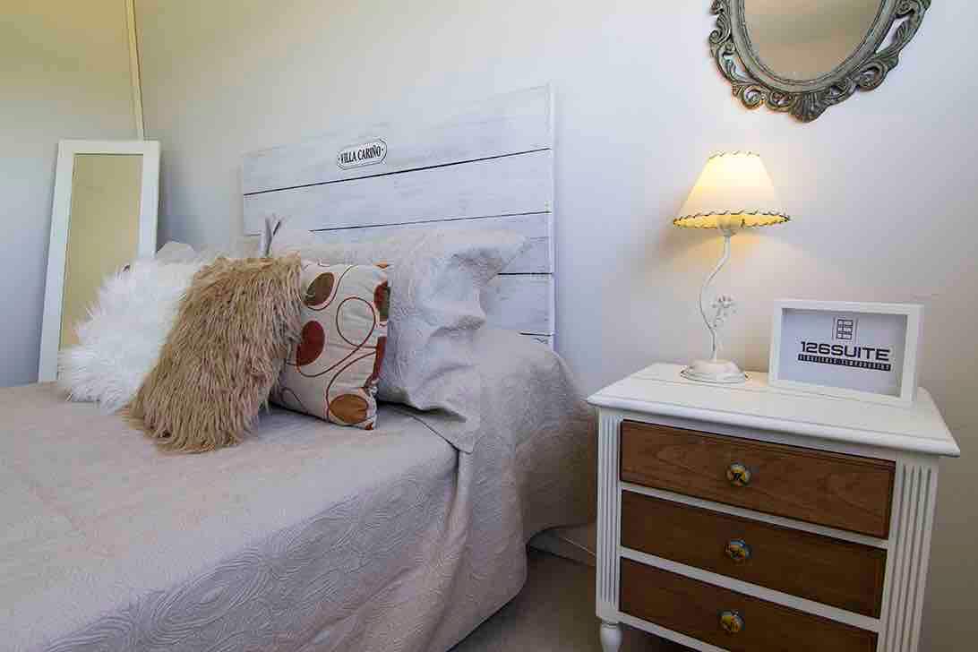A cozy sleeping area features a light-colored headboard with a wooden texture, complemented by a plush bedspread. Decorative pillows add softness, while a bedside lamp provides warm light. A distinctive mirror reflects light, and a small nightstand holds a decorative sign.