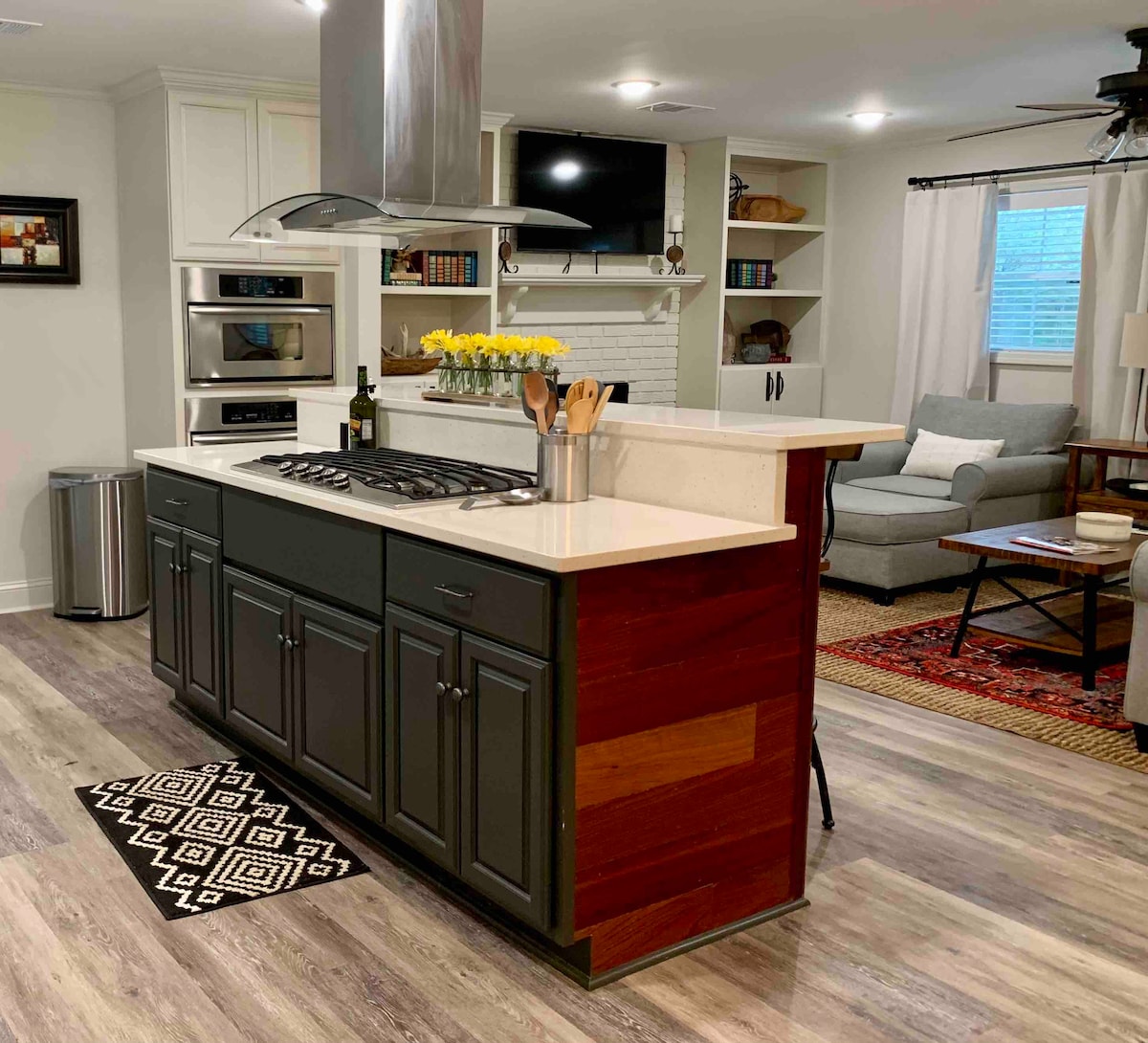 A modern kitchen is displayed, featuring a central island with a gas cooktop. Light-colored cabinets contrast with the dark island cabinetry. Decorative items are arranged on the countertop, and a comfortable living area is visible in the background, showcasing a gray sofa and a bookcase.