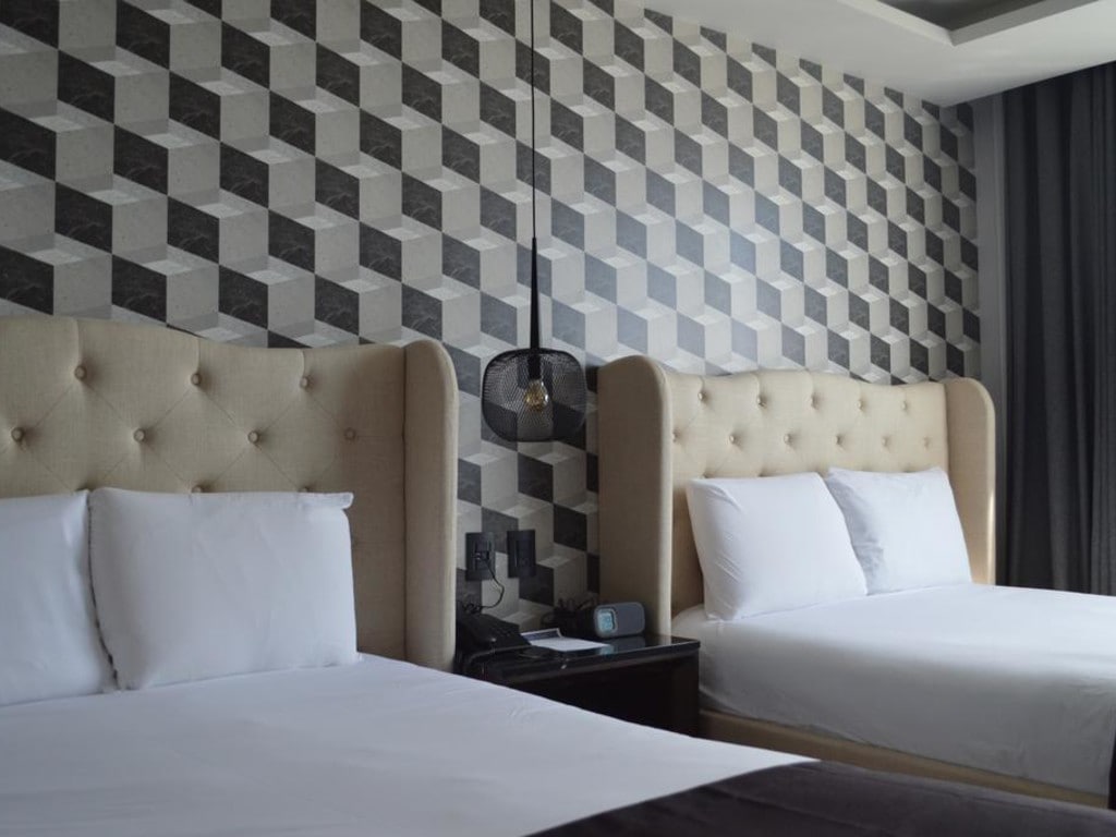 The room features two spacious beds with upholstered headboards against a geometrically patterned accent wall. A bedside table holds a lamp and clock. Nearby, a window is partially covered by dark curtains, allowing natural light to enter the space.