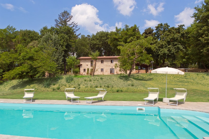 A tranquil swimming pool surrounded by sun loungers and greenery, with a rustic stone villa in the background. The scene is framed by lush trees and a clear blue sky, creating a serene outdoor space perfect for relaxation.