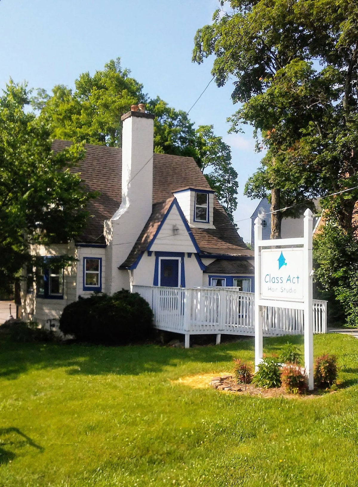 The exterior of a charming two-story building is displayed, featuring white siding with blue accents. A sign labeled 'Class Act Hair Studio' stands at the front, surrounded by well-maintained grass and shrubs. Tall trees provide shade, enhancing the inviting atmosphere of the site.