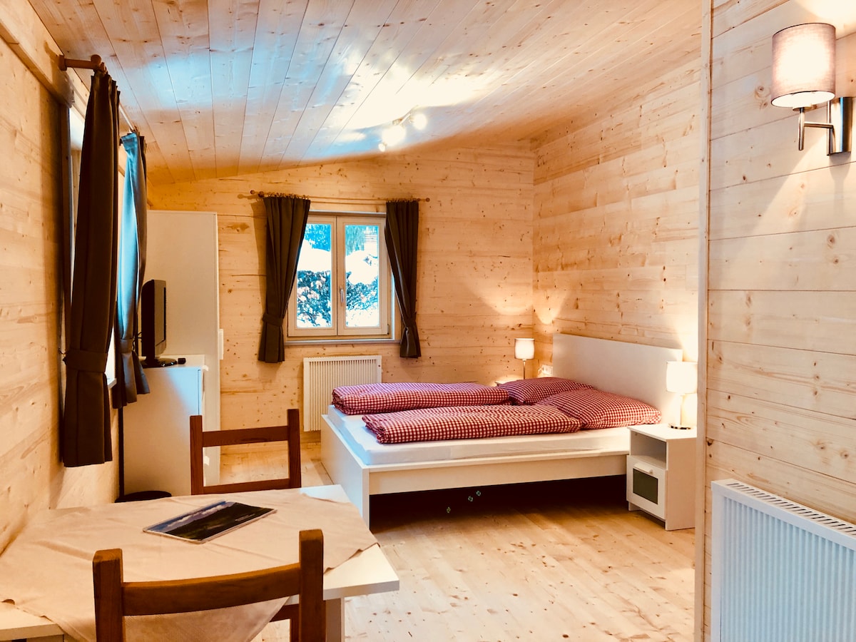 The interior of the chalet features wooden paneling and a light-colored ceiling. A cozy sleeping area contains two lined beds with a red gingham pattern. A small dining table and chairs are positioned in front of a window, which offers a view of the outdoors. Soft lighting is provided by lamps on either side of the beds.