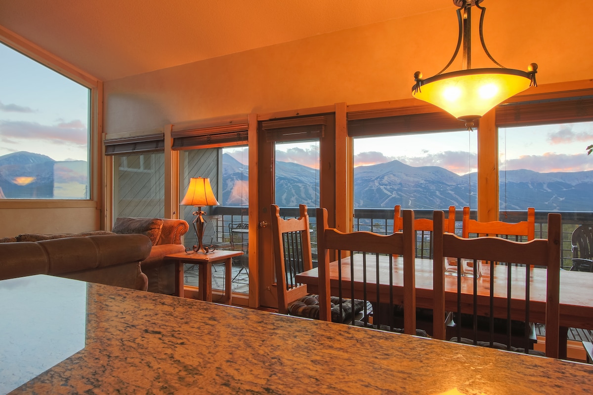 A dining area with a spacious wooden table and chairs is positioned next to large windows, offering expansive views of the mountains. A warm lamp illuminates the space, complementing a cozy sofa visible in the background. The delicate hues of sunset reflect off the window panes.