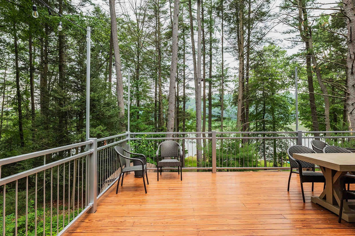 An expansive deck features wooden flooring and a railing, surrounded by tall trees. Comfortable chairs are positioned to offer views of Sweet Arrow Lake, creating a serene outdoor space for relaxation and gatherings.