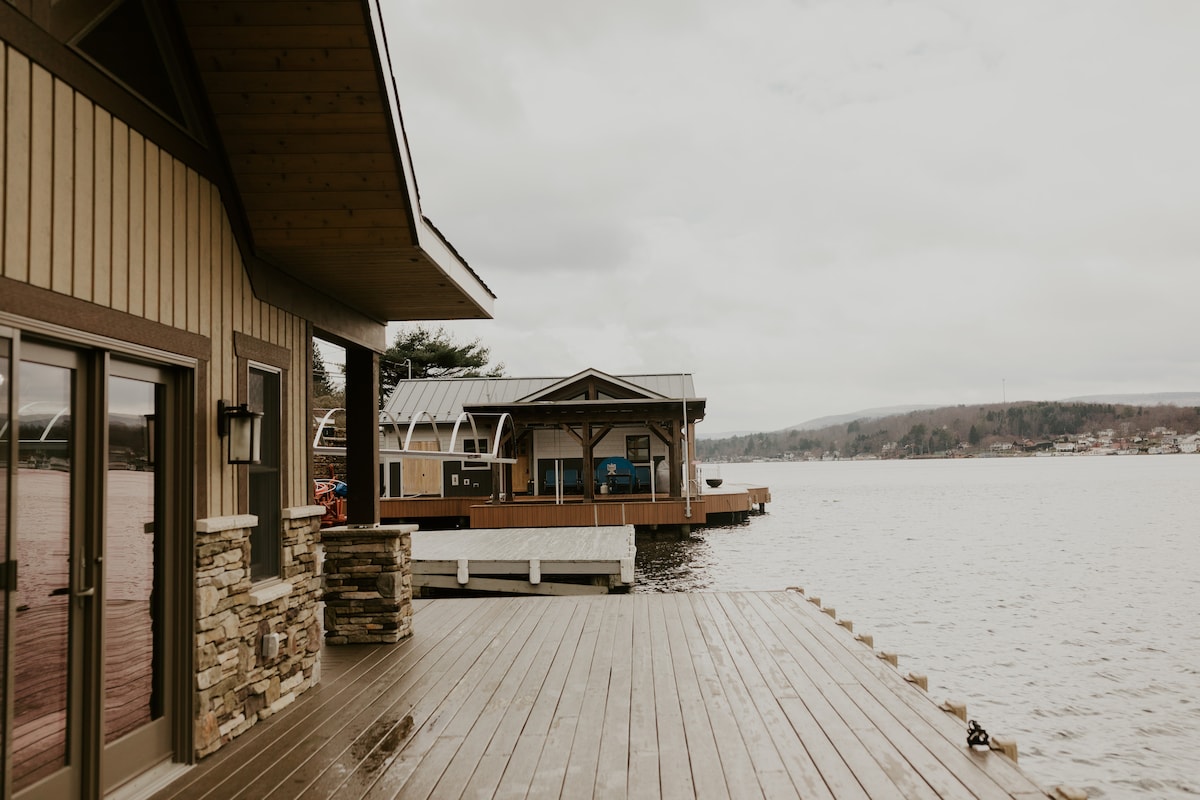 A spacious boardwalk extends alongside the water, connecting to a floating boathouse. The structure features a covered area and is set against a backdrop of gently rolling hills under an overcast sky.