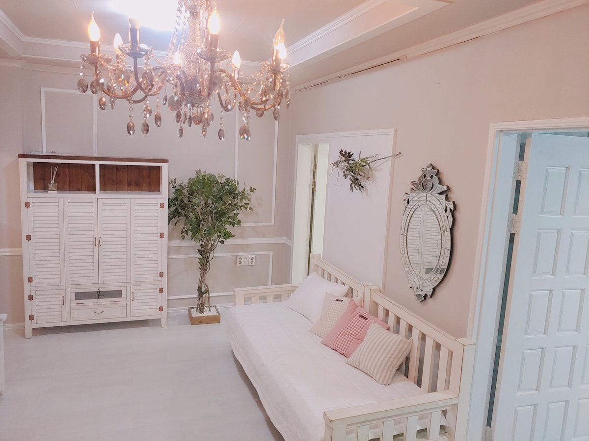 A well-lit living area features a white daybed adorned with light cushions. A vintage-style chandelier hangs above, adding elegance. A decorative mirror and a potted plant enhance the decor, while a white cabinet provides storage. The soft color palette contributes to a calm ambiance.