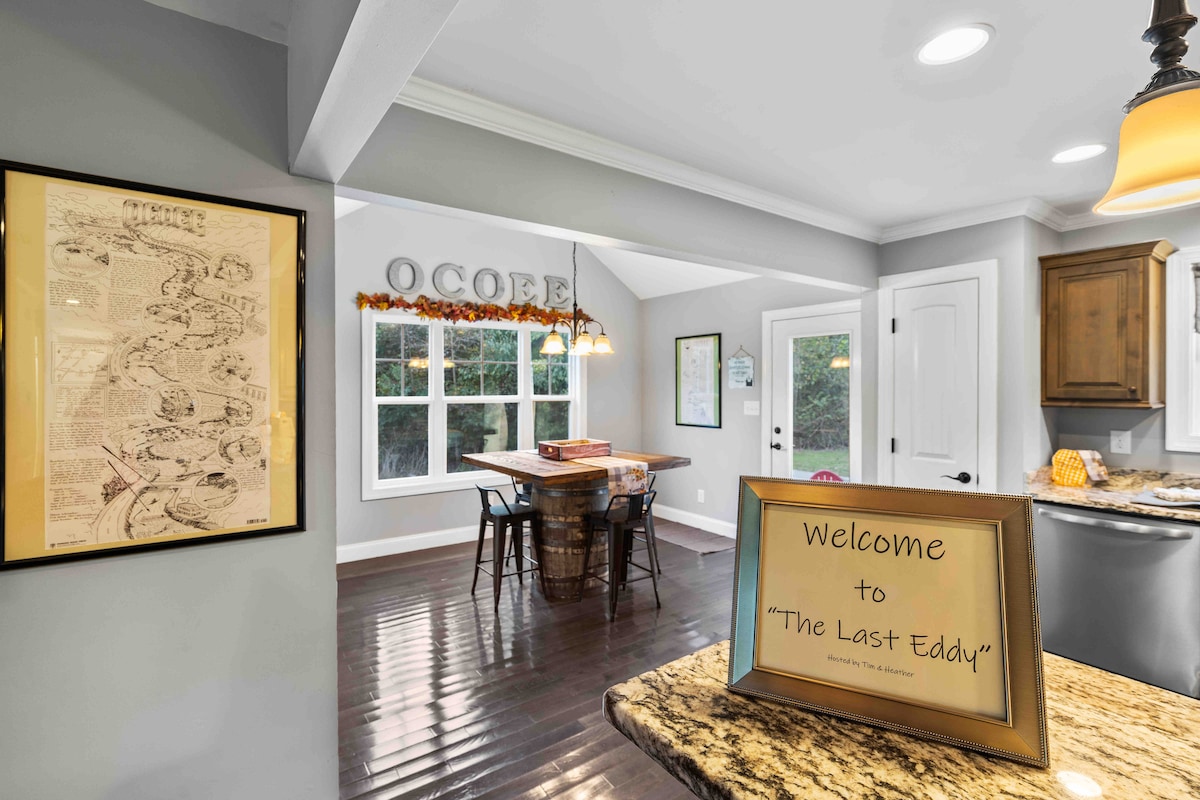 A welcoming entrance includes a framed map on the wall and a sign displaying 'Welcome to The Last Eddy' on the counter. Natural light fills the space through large windows, highlighting a wooden dining table and a rustic barrel serving as a centerpiece.
