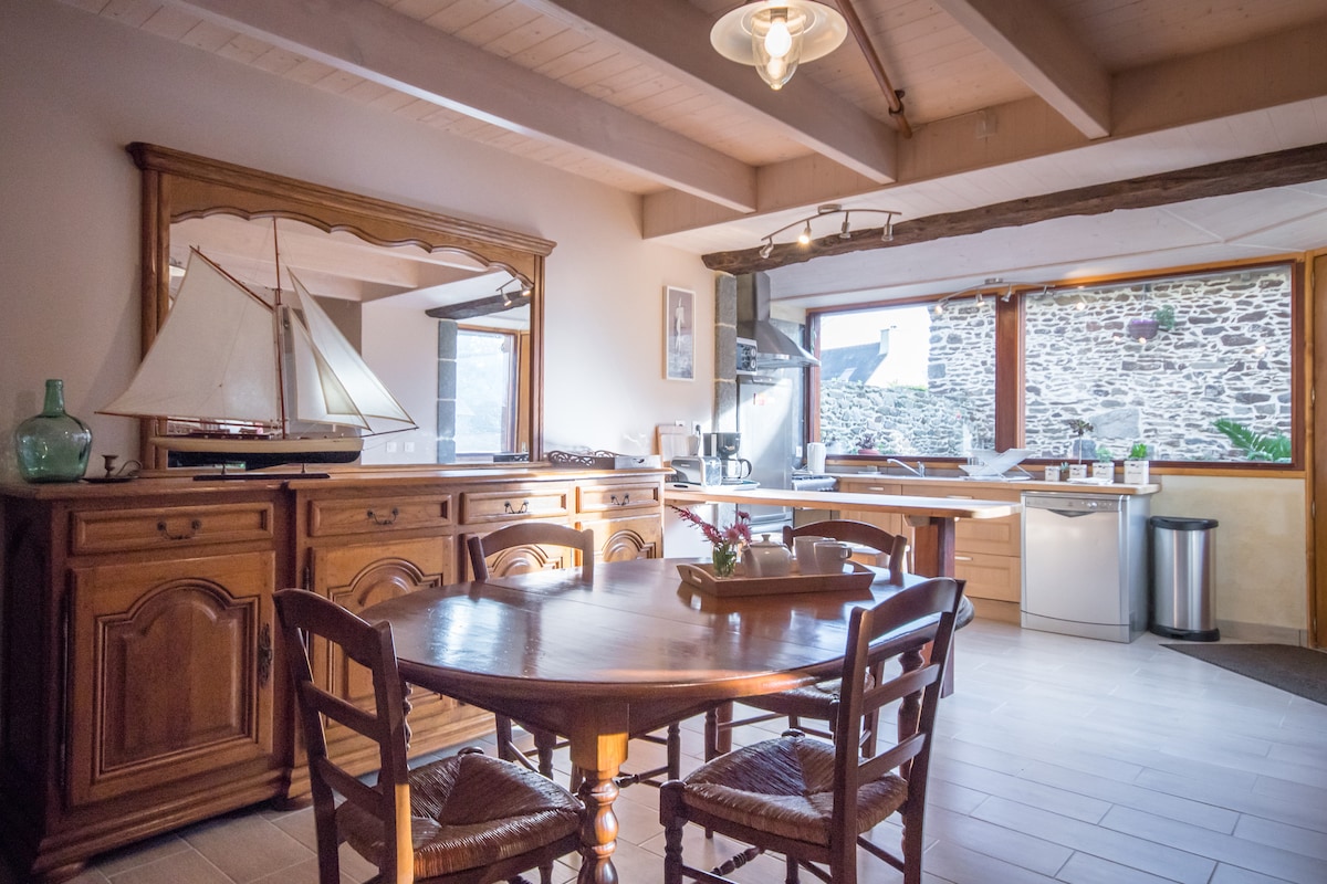 A spacious kitchen area is presented, featuring a wooden dining table surrounded by four woven chairs. A large mirror reflects light, while a detailed model boat decorates the sideboard. Natural light enters through a window, illuminating modern appliances and a rustic stone wall.