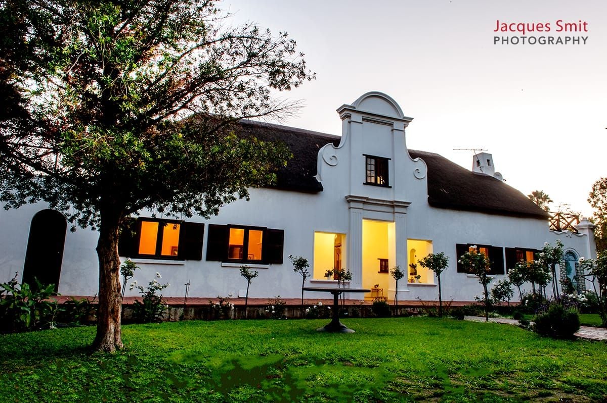 The Cape Dutch style house is set within a lush garden, featuring whitewashed walls and a thatched roof. Soft evening light emanates from the windows, illuminating the surrounding greenery. A small table can be seen on the lawn, nestled among manicured rose bushes.