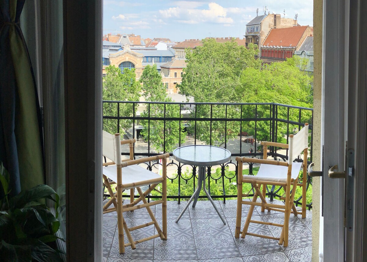 A balcony is visible, featuring two folding chairs and a small round table. The view includes lush green trees and buildings in the background, inviting a serene outdoor setting. The patterned tiles on the balcony floor add a subtle design element.