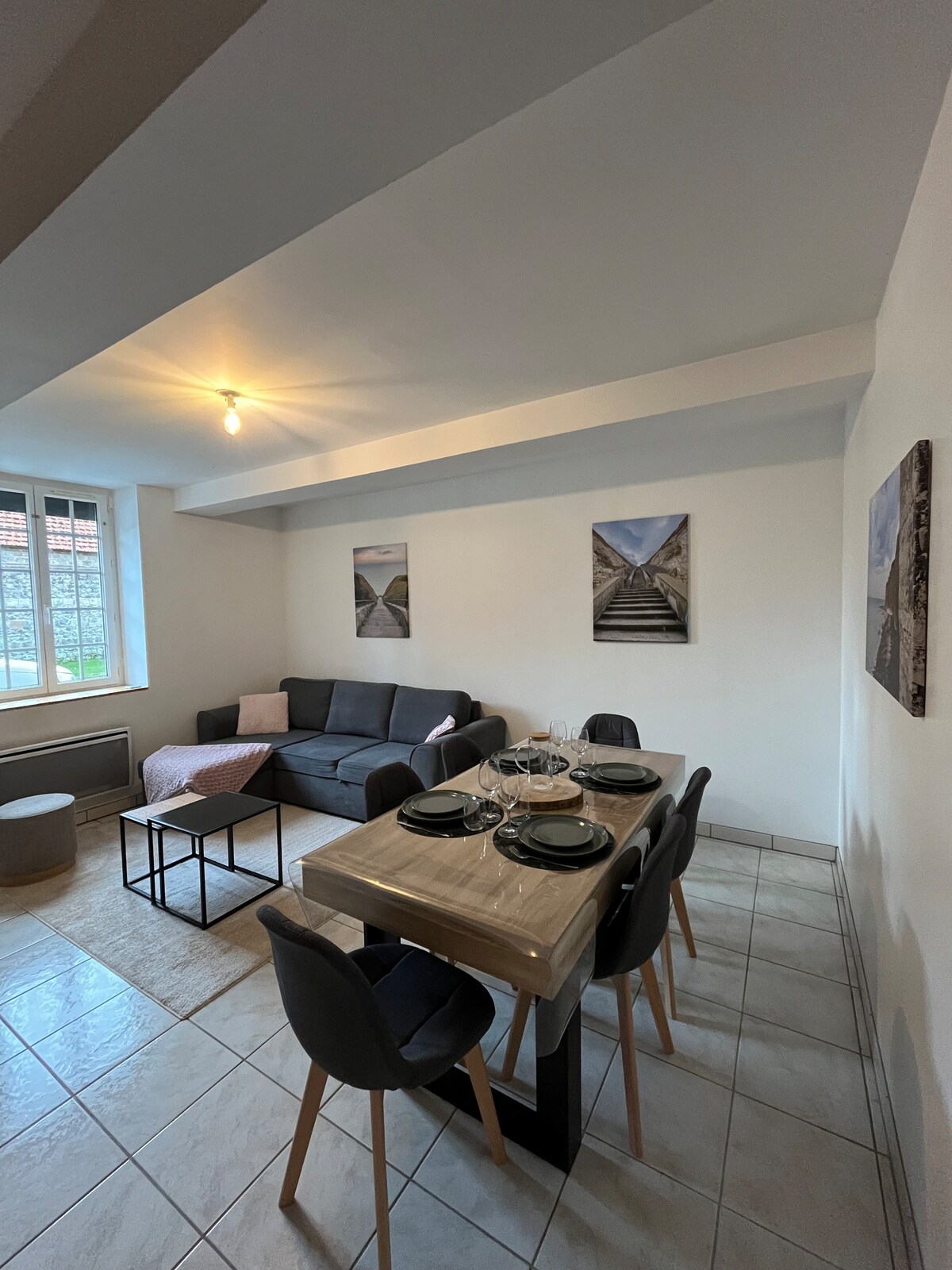 A modern living area is displayed, featuring a comfortable sofa and a dining table set for four. Two framed photographs adorn the walls, enhancing the space. Large windows allow natural light to fill the room, illuminating the neutral-toned interior with tiled flooring.