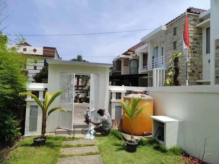 A modern entrance is presented with a white gate and a neatly manicured pathway leading towards the residence. Flanking the path are small plants and an orange water tank. The surrounding buildings exhibit contemporary architectural designs.