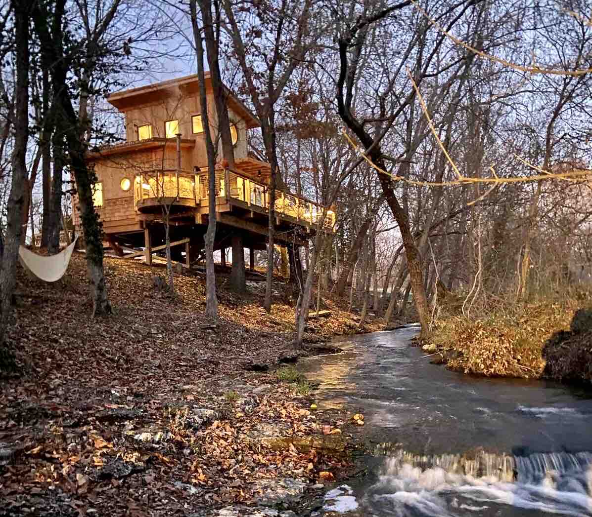 The treehouse sits elevated among trees, showcasing a spacious deck with circular windows. Leaves and twigs cover the ground, while a gentle stream flows nearby. A hammock is visible, inviting relaxation within the serene natural landscape.